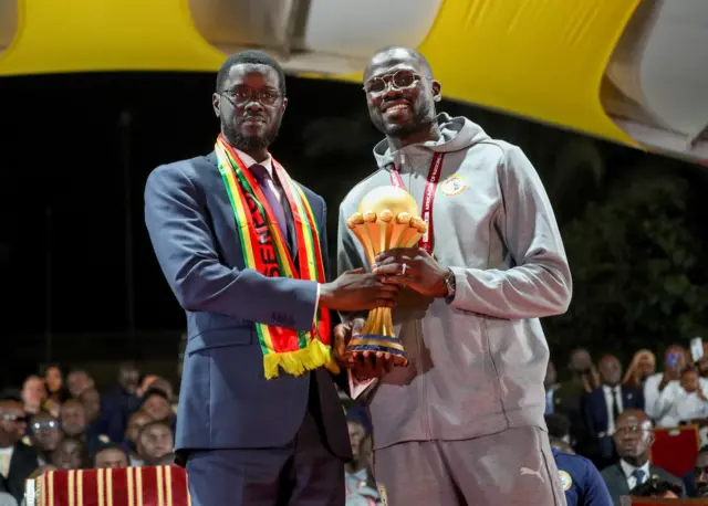 Le président sénégalais Bassirou Diomaye Faye en veste bleue foncée avec une écharpe aux couleurs nationales de son pays, avec le capitaine de l'équipe nationale du Sénégal, Khalidou Koulibaly. Les deux tiennent ensemble dans leurs mains le trophée de la Coupe d'Afrique des Nations.
